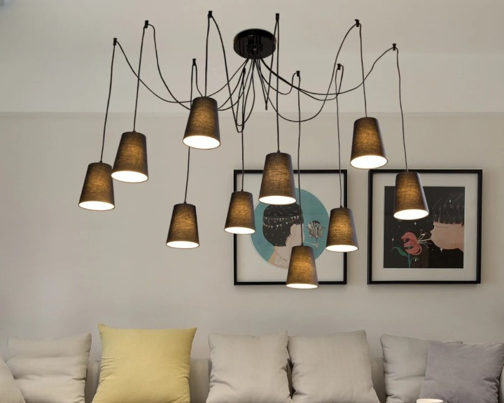 Suspension araignée design et moderne en tissu. A chaque extrémité des câbles, un abat-jour en forme de gobelet à l'envers. Chaque ampoule est allumée. Fixée dans un salon avec un canapé au coussin beige.
