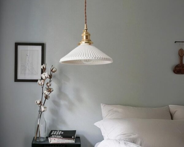 Suspension céramique blanche design épuré, suspendue dans une ambiance cosy de chambre à coucher avec un lit à droite et une petite table de chevet et un cadre à gauche du lit.