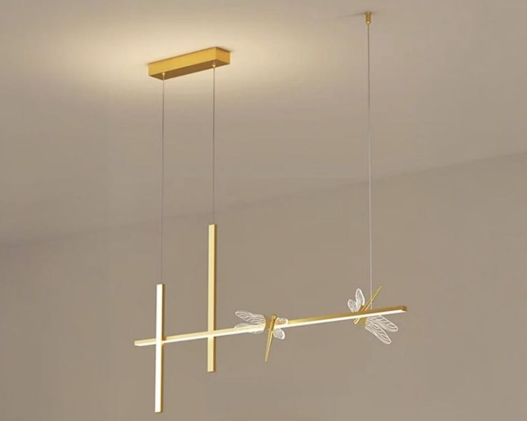 Suspension libellule éclat doré, avec deux libellules, le tout de couleur dorée, luminaire suspendu et allumé. Fond beige.