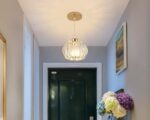 Suspension métal dorée luxueuse allumée dans un couloir avec un bouquet de fleurs sur la droite et une porte au fond.