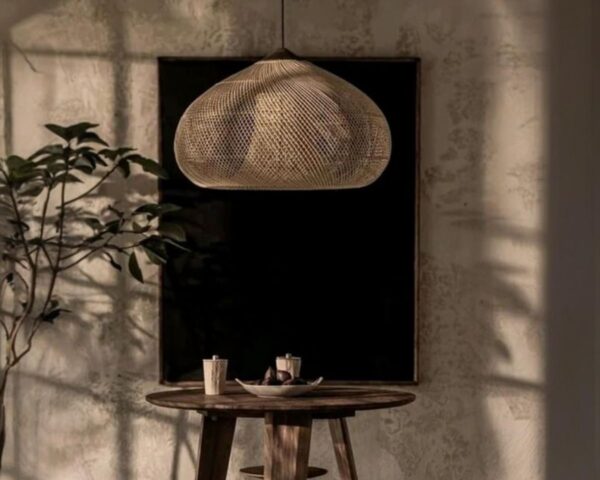 Suspension osier design épuré, de couleur beige, suspendue au-dessus d'une petite table de salon en bois et avec un fond rectangulaire noir sur un mur beige.