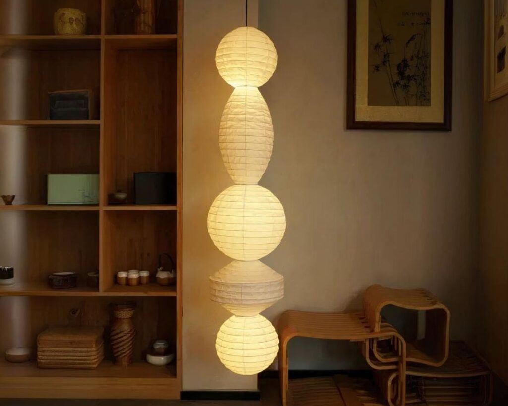 Suspension papier japonais à multiformes allumée dans une pièce avec une étagère en bois sur la gauche.