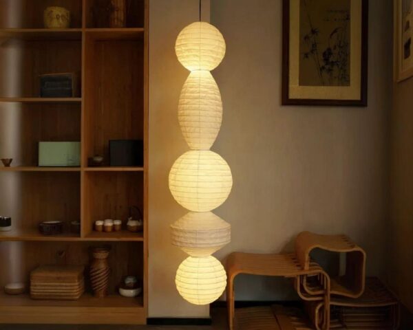 Suspension papier japonais à multiformes allumée dans une pièce avec une étagère en bois sur la gauche.