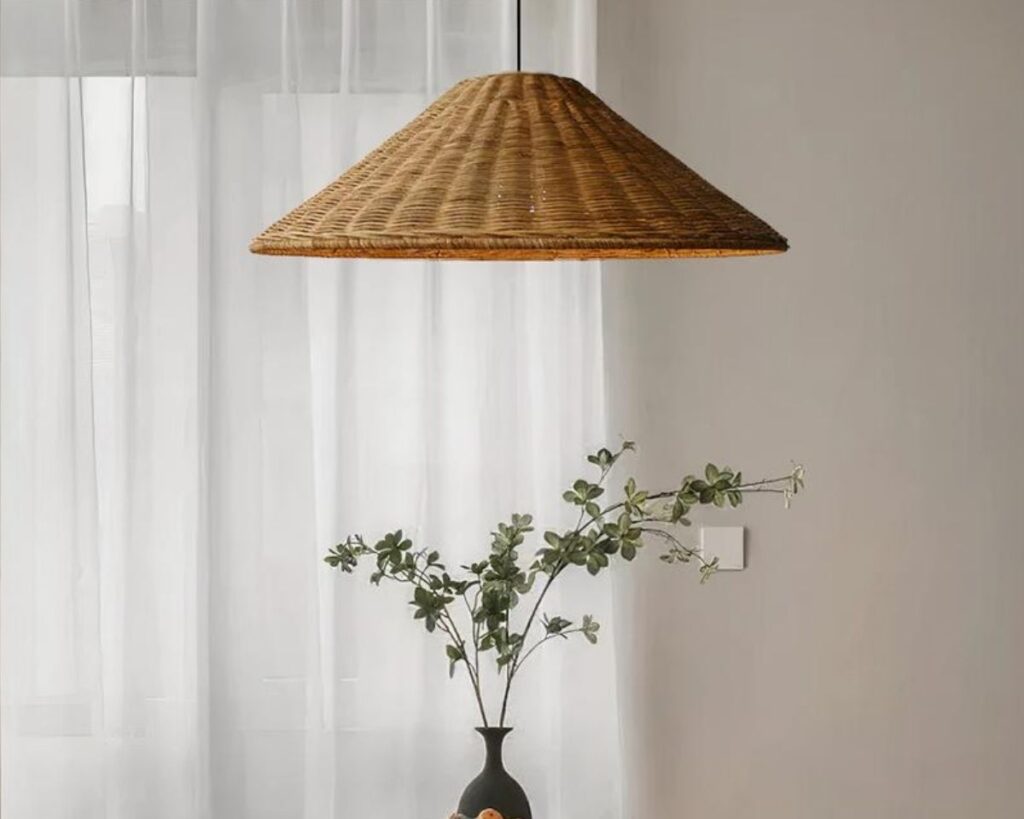 Suspension rotin marron en forme de chapeau au-dessus d'un vase noir avec des branchages verts. En fond, un rideau blanc.