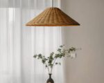 Suspension rotin marron en forme de chapeau au-dessus d'un vase noir avec des branchages verts. En fond, un rideau blanc.