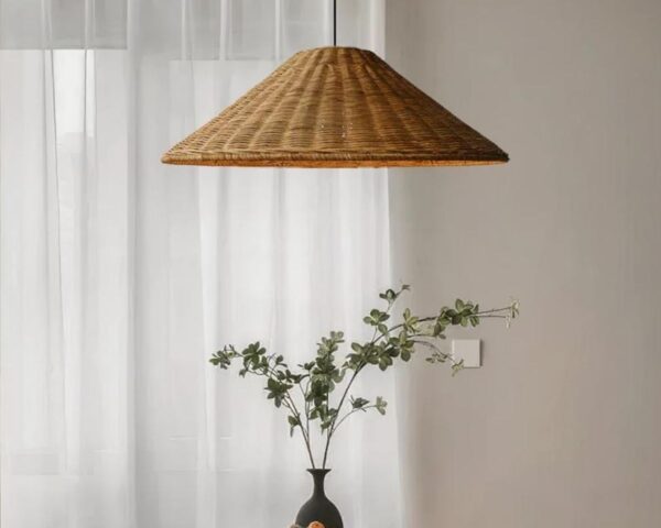 Suspension rotin marron en forme de chapeau au-dessus d'un vase noir avec des branchages verts. En fond, un rideau blanc.