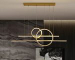 Suspension XXL art moderne dorée, avec deux cercles et deux barres latéral, allumée, suspendue au-dessus d'une table et dans un décor de salon.