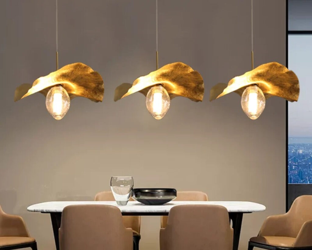 Suspension salon dorée style feuille de lotus allumée au nombe de trois au-dessus d'une table avec 4 chaises autour. Le mur en fond est beige.