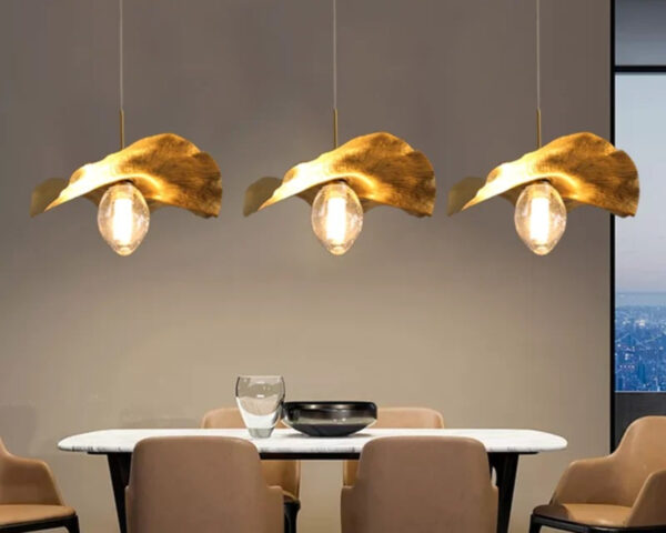 Suspension salon dorée style feuille de lotus allumée au nombe de trois au-dessus d'une table avec 4 chaises autour. Le mur en fond est beige.