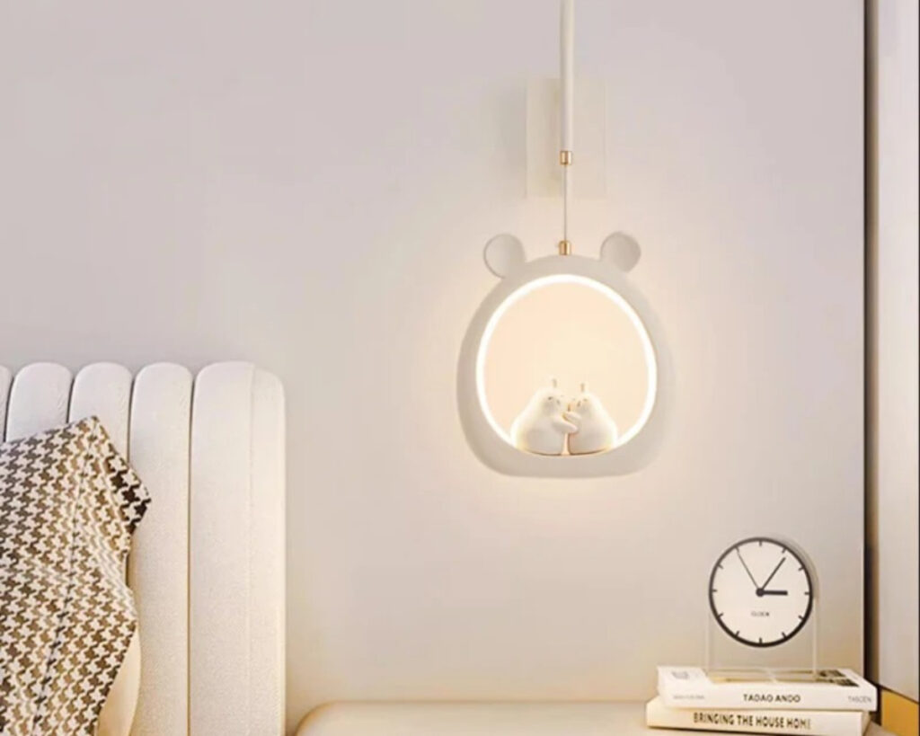 Suspension chambre bébé décorative à lumière variable en forme de cercle blanc avec des oreilles d'ourson sur le dessus. Le cercle est éclairé avec deux personnages au centre. Un lit à gauche et une table de chevet avec un réveil à droite.