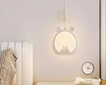 Suspension chambre bébé décorative à lumière variable en forme de cercle blanc avec des oreilles d'ourson sur le dessus. Le cercle est éclairé avec deux personnages au centre. Un lit à gauche et une table de chevet avec un réveil à droite.