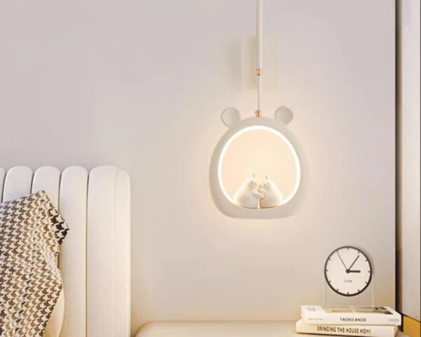 Suspension chambre bébé décorative à lumière variable en forme de cercle blanc avec des oreilles d'ourson sur le dessus. Le cercle est éclairé avec deux personnages au centre. Un lit à gauche et une table de chevet avec un réveil à droite.