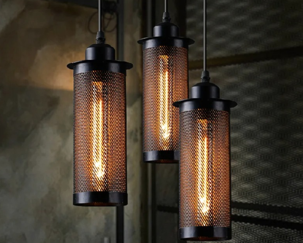 Suspension industrielle au design en maille de fer au nombre de trois et allumées. En arrière plan, un mur gris et une grille sur la droite.