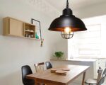 Suspension cuivre style vintage de couleur noire avec une ampoule allumée fixée dans une pièce au-dessus d'une table rectangulaire. A gauche, un mur blanc avec un petit meuble beige. A droite, un petit comptoir avec une petite plante verte.