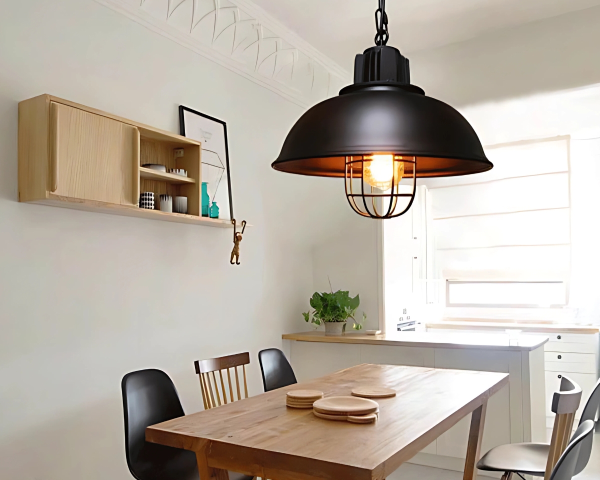 Suspension cuivre style vintage de couleur noire avec une ampoule allumée fixée dans une pièce au-dessus d'une table rectangulaire. A gauche, un mur blanc avec un petit meuble beige. A droite, un petit comptoir avec une petite plante verte.
