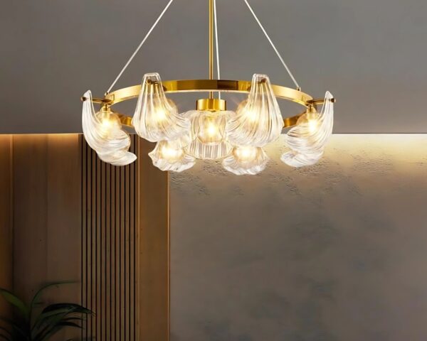 Suspension coquillage 9 têtes sur un anneau doré suspendu. Les ampoules sont allumées. Le mur à l'arrière est beige avec la partie de gauche marron style bois. Une plante verte en bas à gauche.