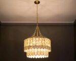 Suspension coquillage luxueuse en cuivre allumée. Elle est composée de deux niveaux de coquillages placés en forme de rond sur une structure dorée. Fixée sur un plafond blanc et le mur à l'arrière est marron.