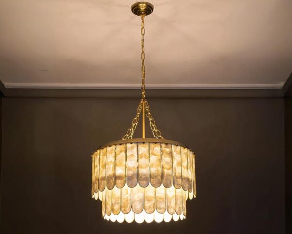Suspension coquillage luxueuse en cuivre allumée. Elle est composée de deux niveaux de coquillages placés en forme de rond sur une structure dorée. Fixée sur un plafond blanc et le mur à l'arrière est marron.