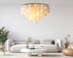 Suspension coquillage style chandelier allumée. Fixée dans un salon avec un canapé moderne gris clair, une table basse blanche, une plante verte sur la gauche du canapé et un fauteuil marron à droite.