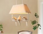 Suspension japonaise avec trois oiseaux blancs posés sur une barre au-dessus de laquelle il y a un abat-jour faisant office de toit. La suspension est allumée dans une pièce au mur beige avec une chaise et des branchages à droite.