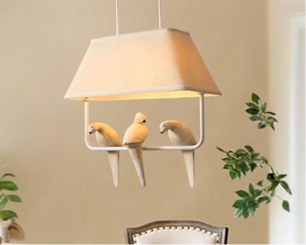 Suspension japonaise avec trois oiseaux blancs posés sur une barre au-dessus de laquelle il y a un abat-jour faisant office de toit. La suspension est allumée dans une pièce au mur beige avec une chaise et des branchages à droite.
