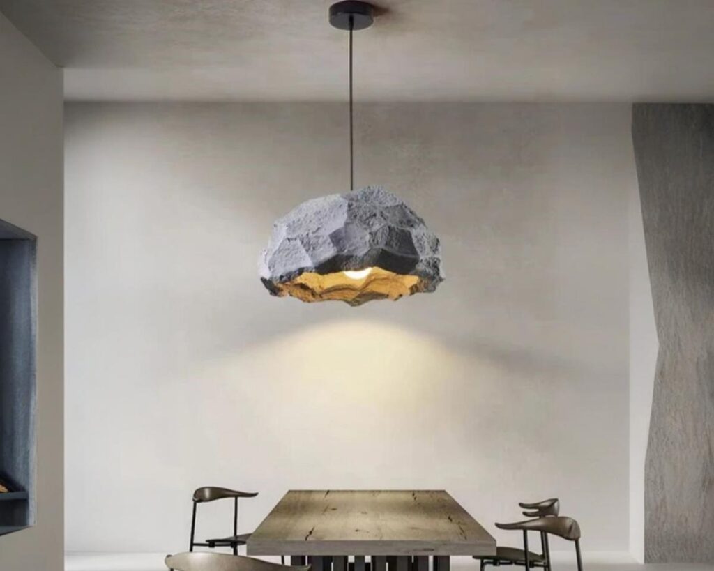 Suspension LED en forme de roche, de couler grise, allumée et suspendue au-dessus d'une table. Fond beige foncé.