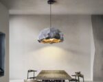 Suspension LED en forme de roche, de couler grise, allumée et suspendue au-dessus d'une table. Fond beige foncé.
