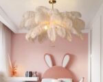 Suspension plume design feuilles de couleur blanche avec une barre de suspension dorée. L'ampoule est allumée. Fixée dans une chambre d'enfant au mur rose avec un lit rose à oreilles de lapin.