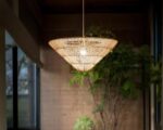 Suspension en rotin beige en forme de cône inversé, dans une pièce avec des plantes en fond.