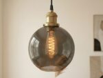 La suspension industrielle design chic en verre fumé avec douille laiton doré et ampoule filament, au-dessus d'une table chêne.