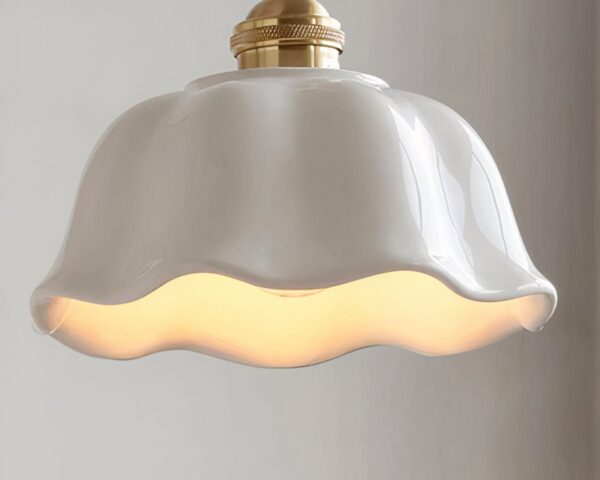 Une suspension en céramique blanche à bord ondulé campagne chic, avec abat-jour blanc brillant et lumière chaude, suspendue devant un mur clair.