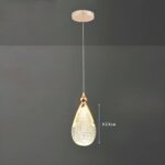 Une suspension goutte d’eau en acrylique doré avec un abat-jour en forme de goutte brillante, suspendue à un long fil sur fond gris.
