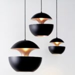 Trois suspension sphérique design à double coque blanc et doré, avec abat-jour noirs suspendus sur fond clair, diffusant une lumière chaude.