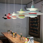 Une suspension design danois à abat-jour multicouches colorée, suspendue au-dessus d’une table en bois dans une salle à manger simple et lumineuse.